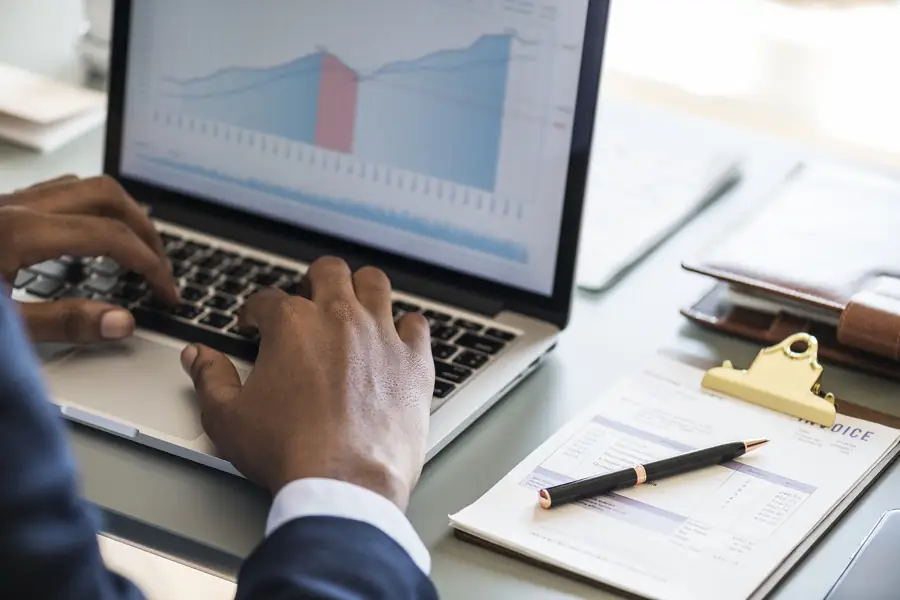 Hands on a laptop keyboard beside charts and an invoice on a clipboard